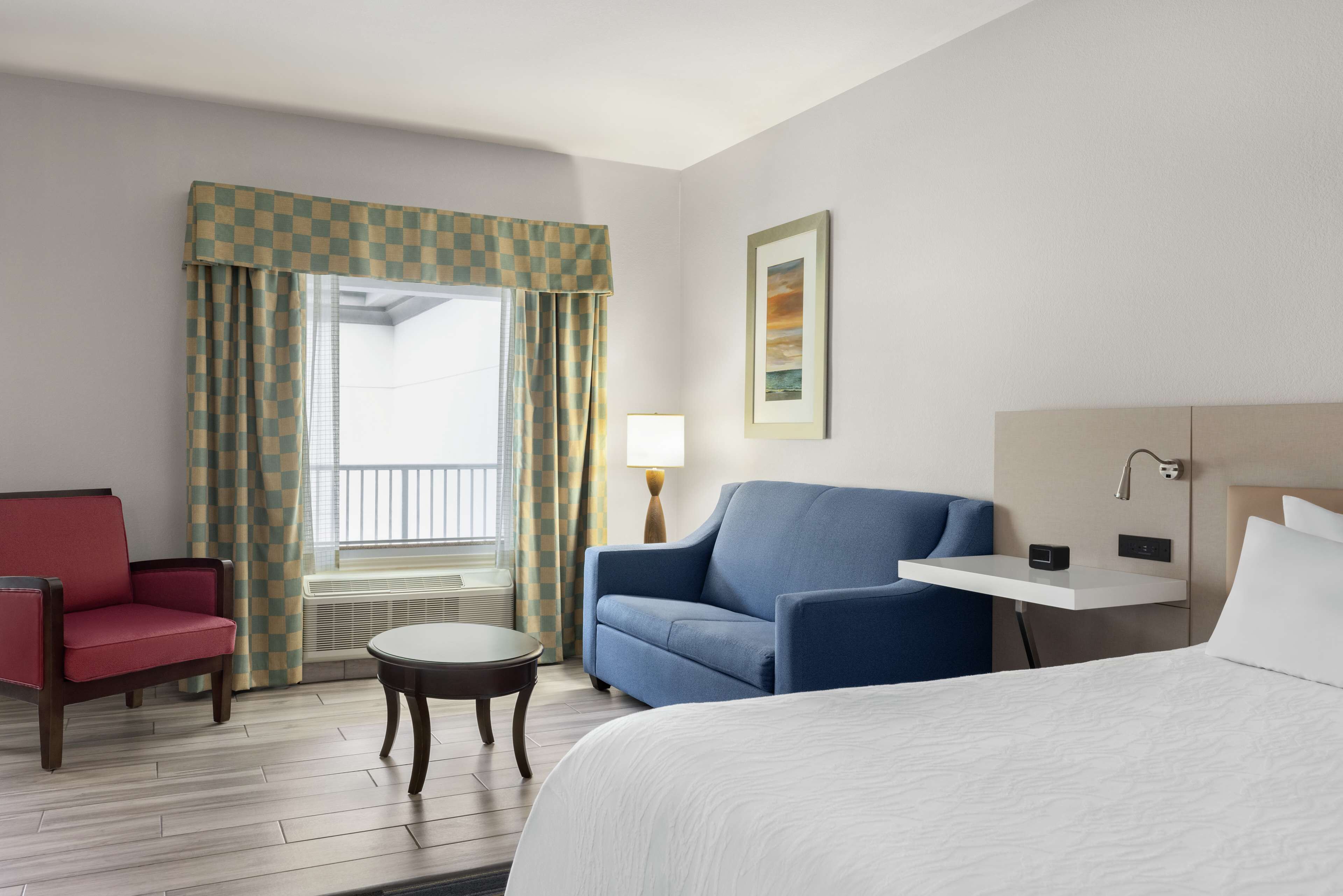 Una camera d'albergo con un letto, una poltrona blu, una poltrona rossa, un tavolino rotondo, una lampada, un quadro appeso e una finestra con tende a quadretti.