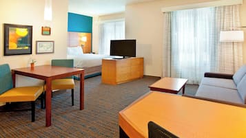 A hotel room with a bed, a TV on a wooden dresser, a sofa, a dining table with chairs, and a small table with a lamp.