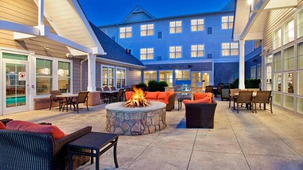 An outdoor patio with stone fire pit, seating, and a view of a large building in the background. - Residence Inn Bridgewater Branchburg (Branchburg)