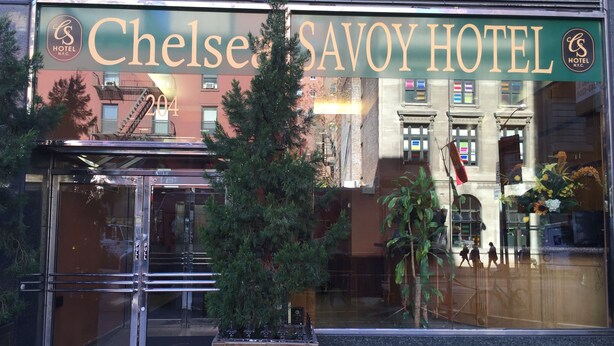 The Chelsea Savoy Hotel entrance with a glass door, a potted tree, and a reflection of the building's facade. - Chelsea Savoy Hotel (New York)
