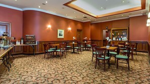 A hotel dining area with a patterned carpet, wooden tables and chairs, and wall-mounted light fixtures. - Chelsea Savoy Hotel (New York)