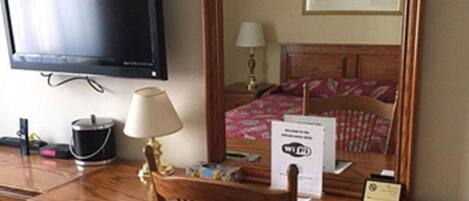 A hotel room with a wooden desk, a television, a lamp, a bed with a patterned bedspread, and a framed picture on the wall.