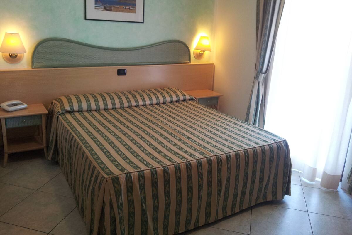 A hotel room with a bed, two bedside tables, a nightstand, a wall-mounted picture, and two wall lamps.