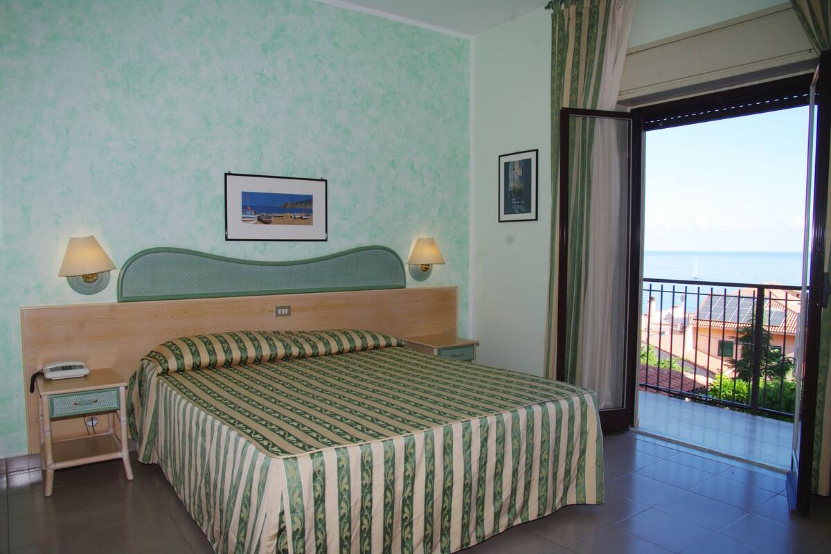 A hotel room with a bed, two bedside lamps, a nightstand, a window with a view of the sea, and a door leading to an outdoor balcony.