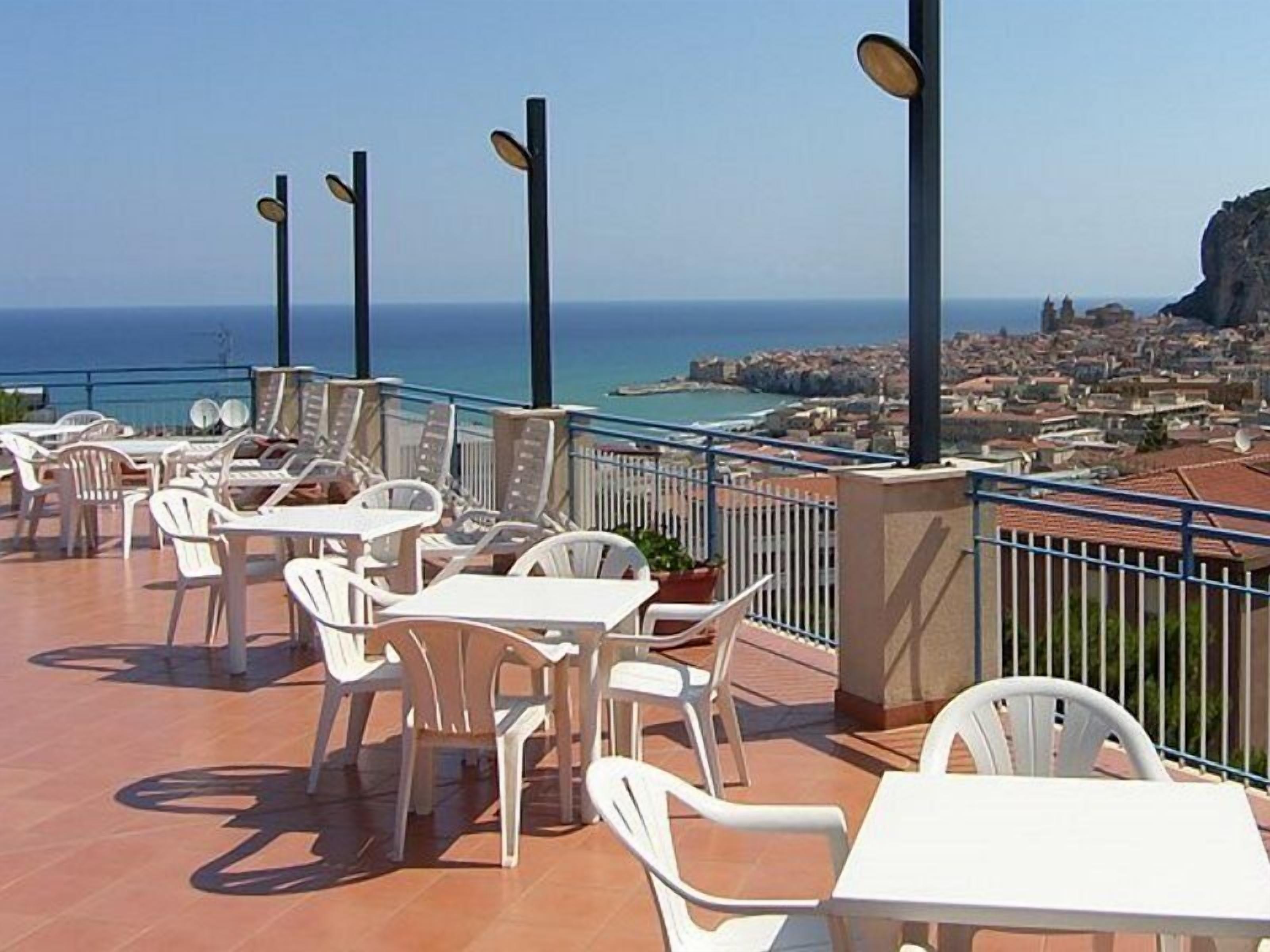 Una terraza al aire libre con sillas y mesas de plástico blanco, baranda y vistas al mar y al paisaje urbano.