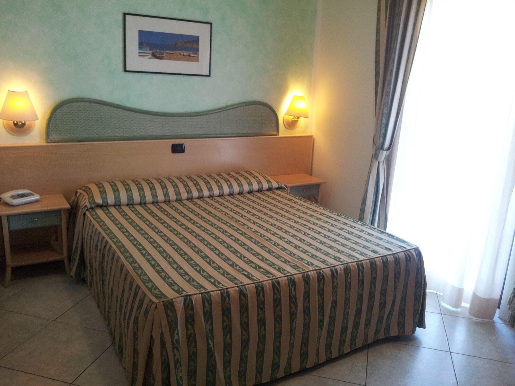 Une chambre d’hôtel avec un lit, deux tables de chevet, une commode, une lampe, un tableau au mur et une fenêtre avec des rideaux.
