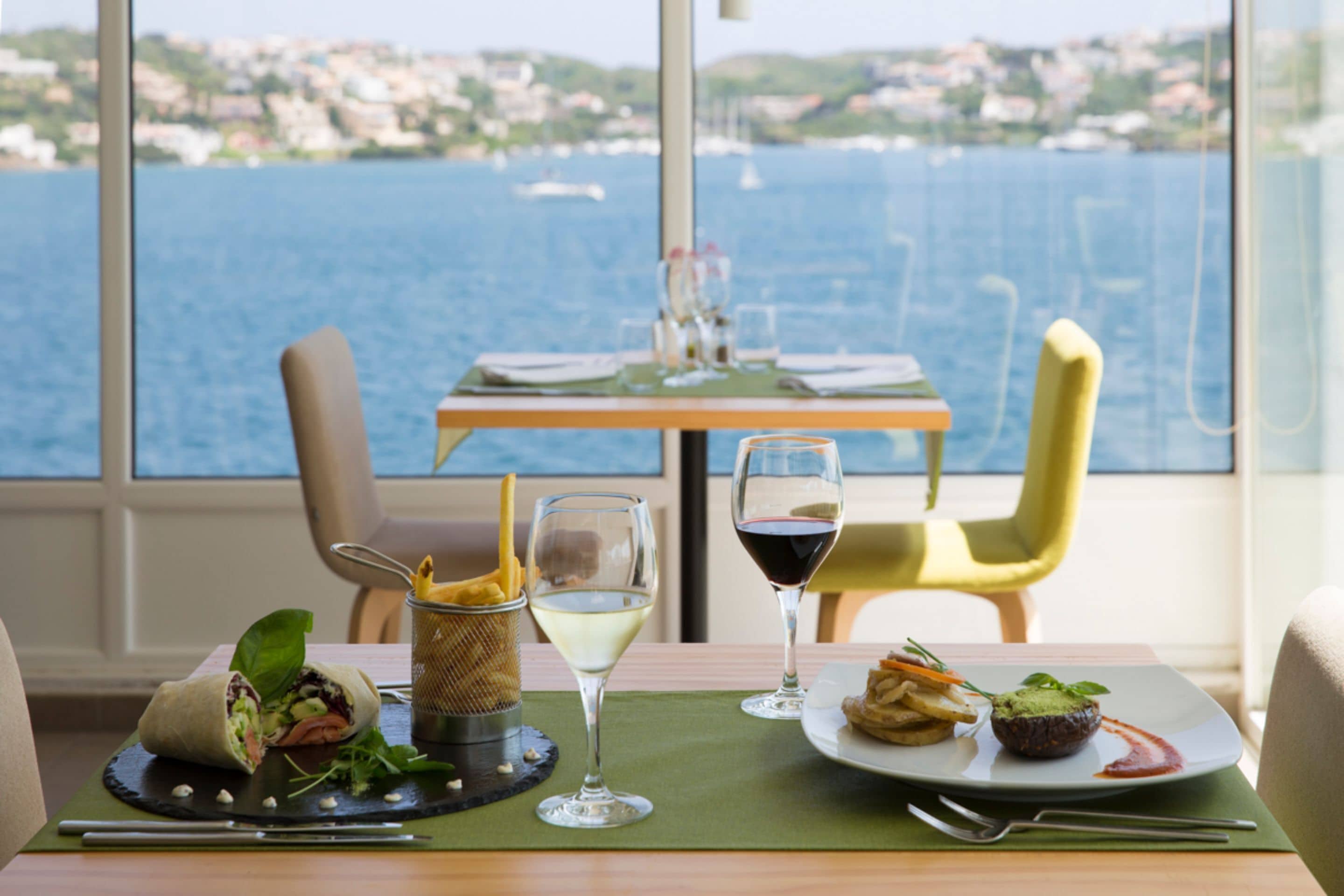 A dining table with a meal, a glass of white wine, and a glass of red wine. The table is set with a green placemat, and there is a view of the sea and buildings in the background.