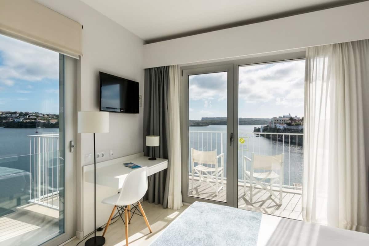 Habitación de hotel moderna con balcón, escritorio, silla, televisión y vistas al agua y edificios.