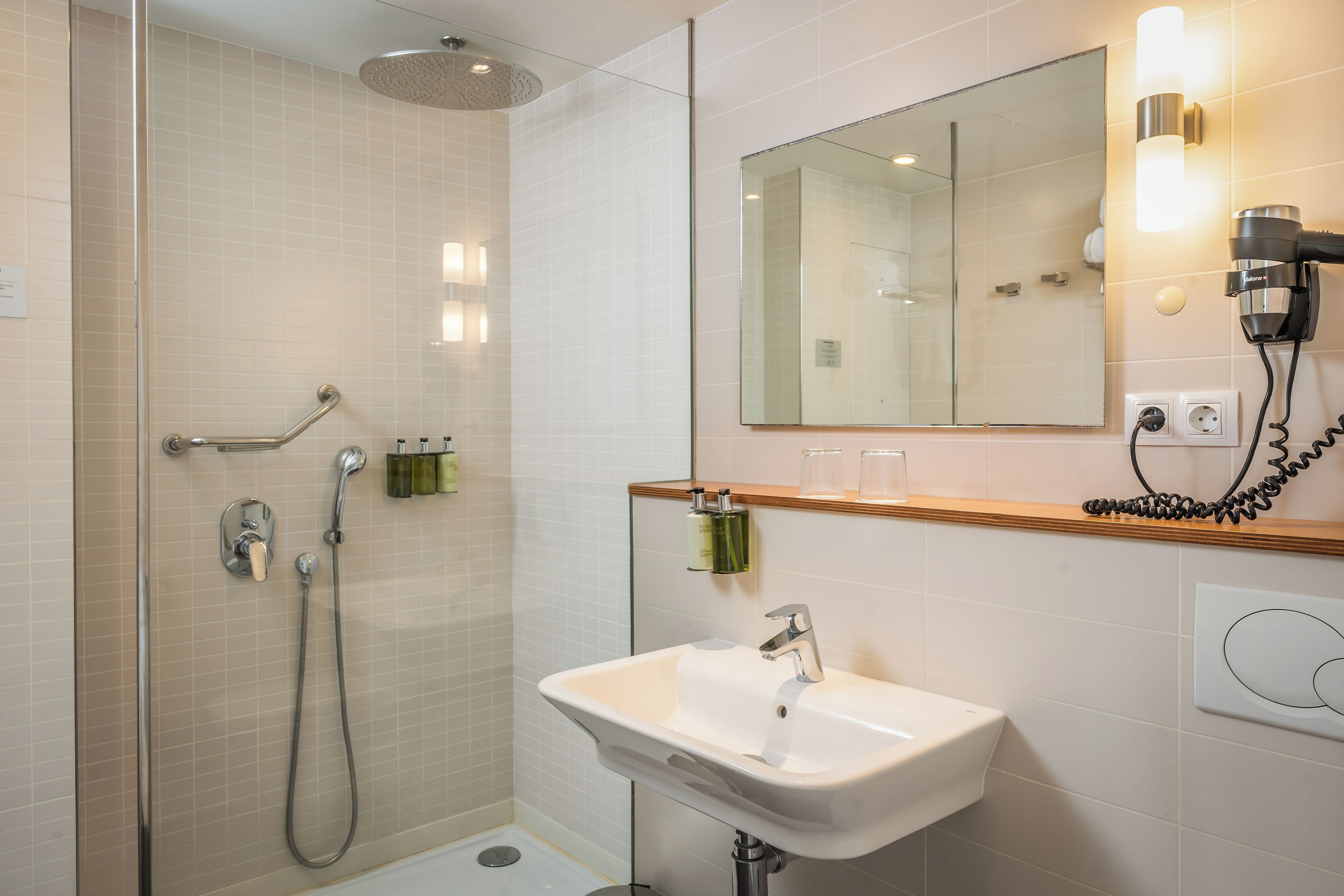 Un baño moderno con ducha envuelta en vidrio, un lavamanos con espejo y un secador de pelo en la pared.