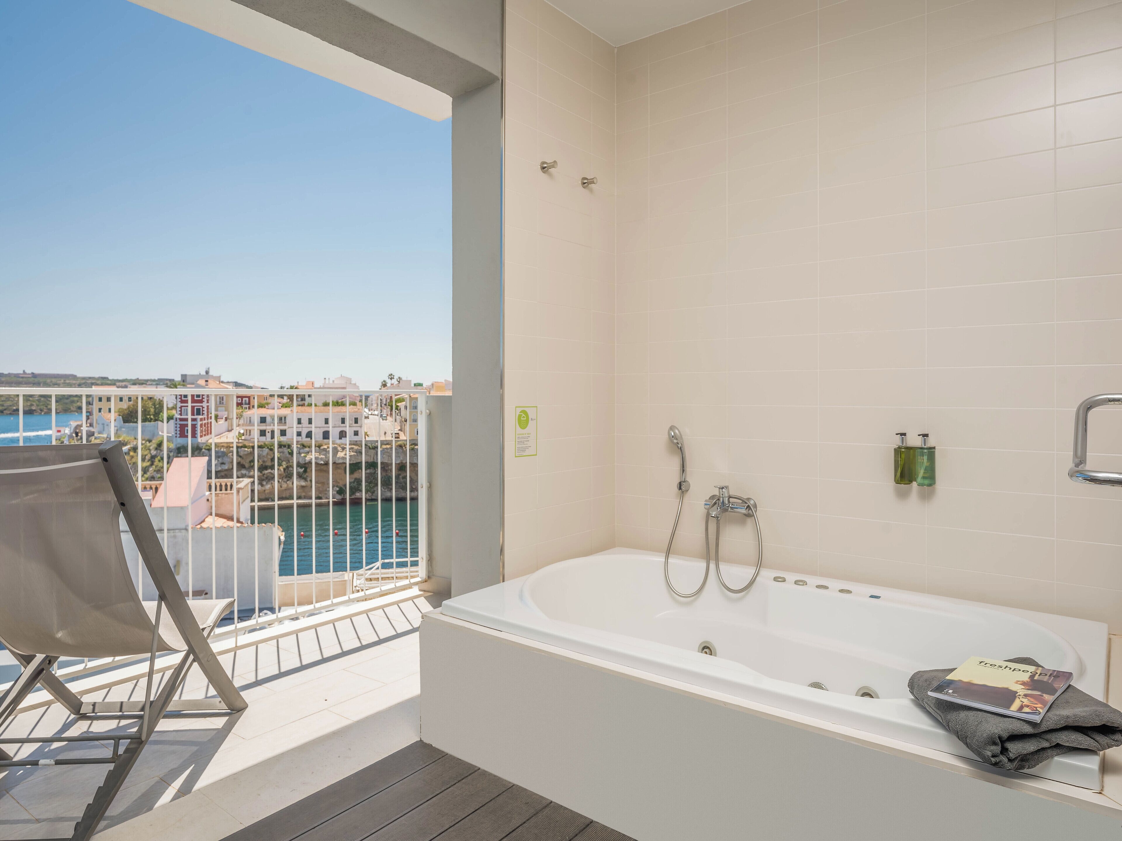 Un balcón con vista al mar y edificios, una bañera blanca con un libro y una toalla.