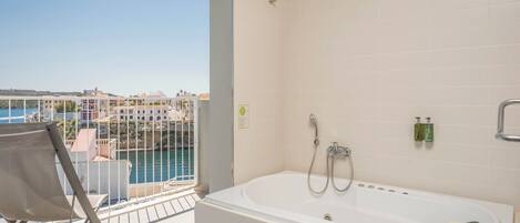 A balcony with a view of the sea and buildings, a white bathtub with a book, and a towel.