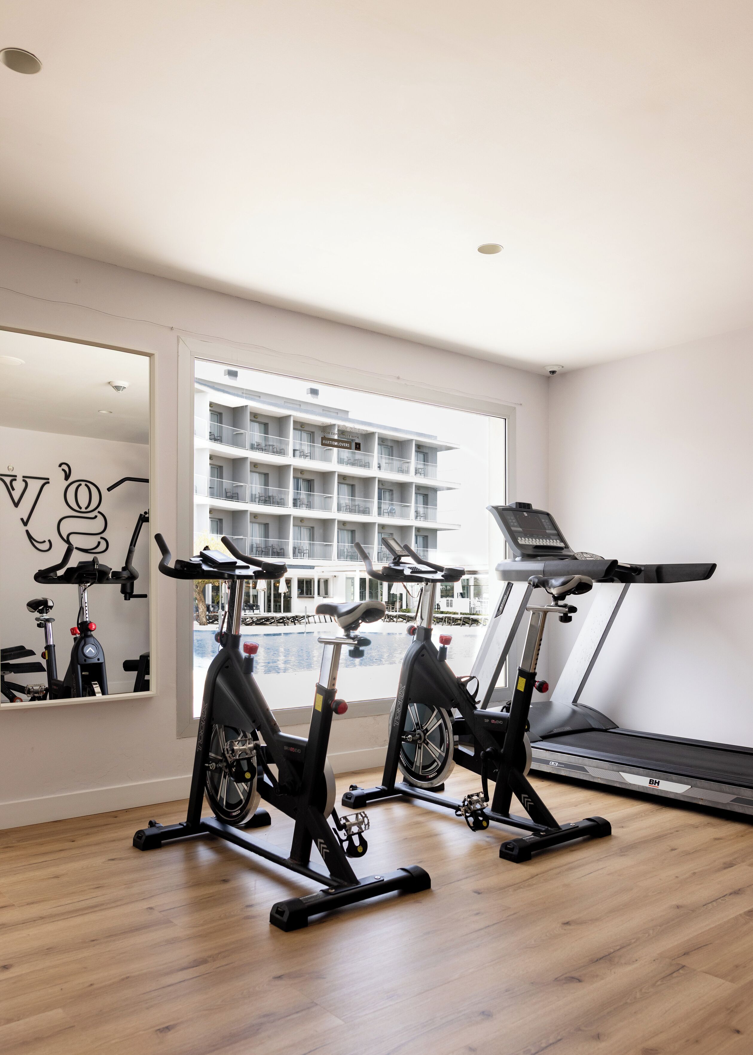 A gym with two stationary bikes and a treadmill, large windows, and a view of a building with balconies.