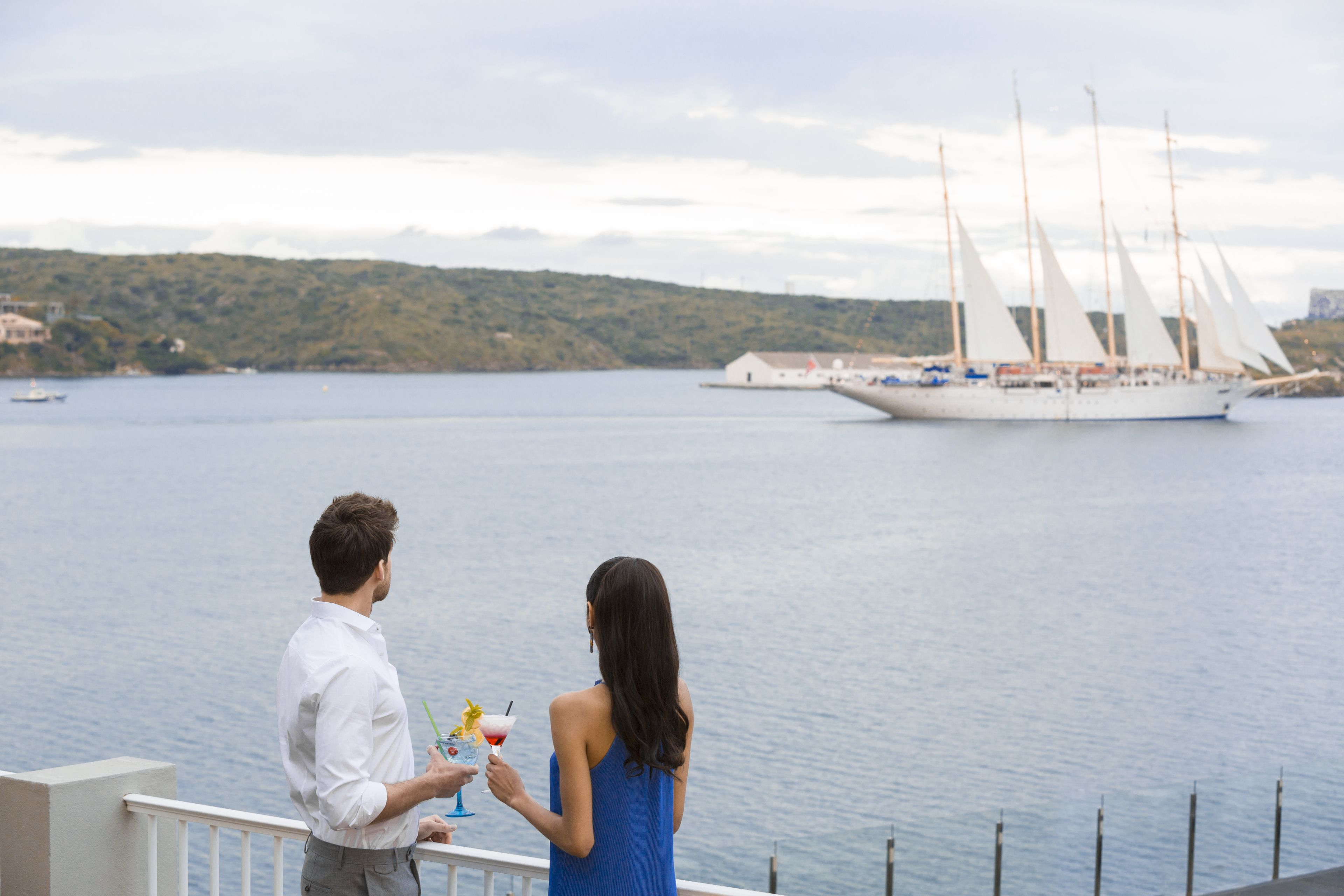 Una pareja parada junto a un balcón con vista a un cuerpo de agua y un velero a lo lejos.
