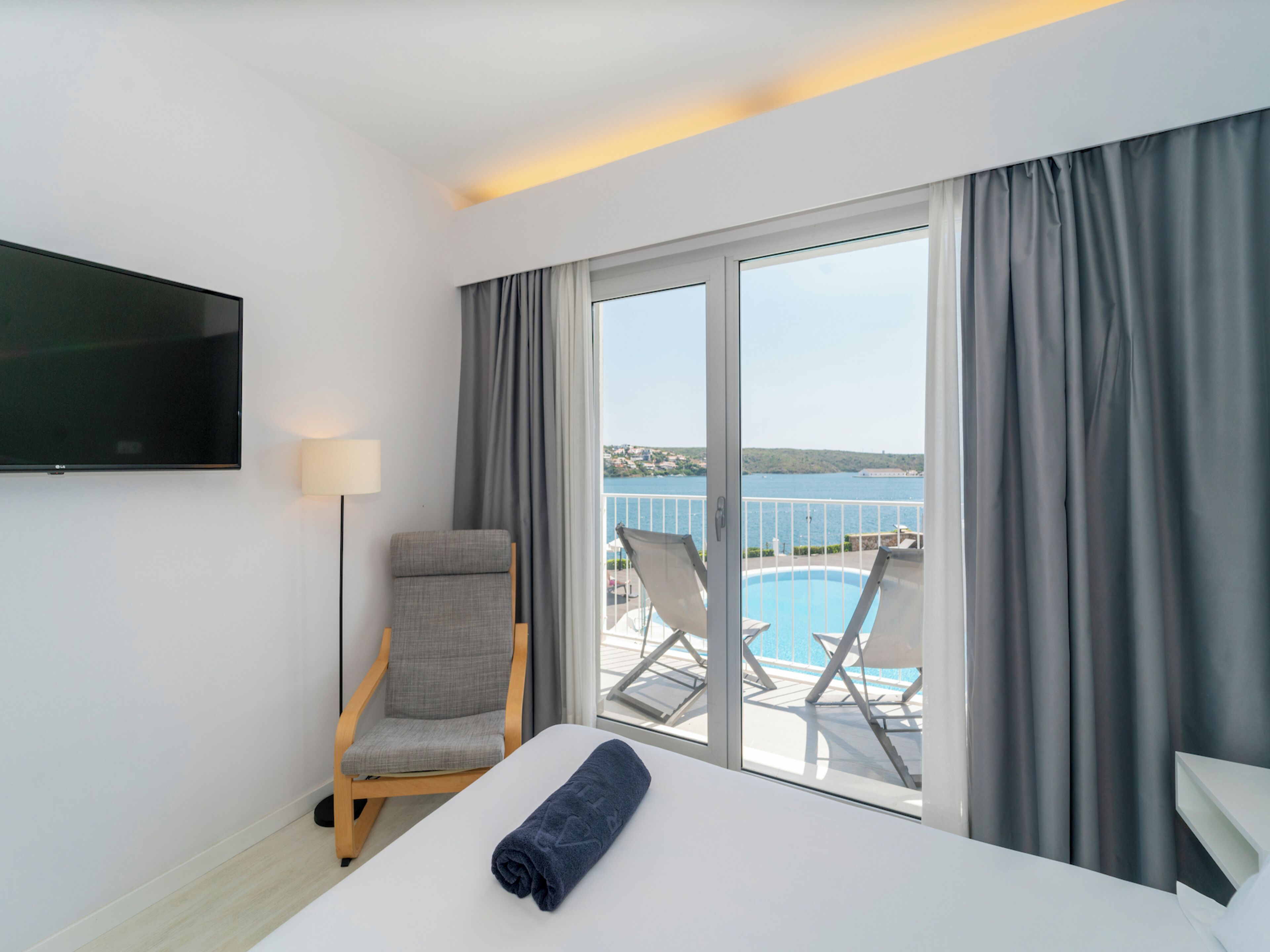 Habitación de hotel con cama, silla, televisor, lámpara y vista a una piscina y paisaje.
