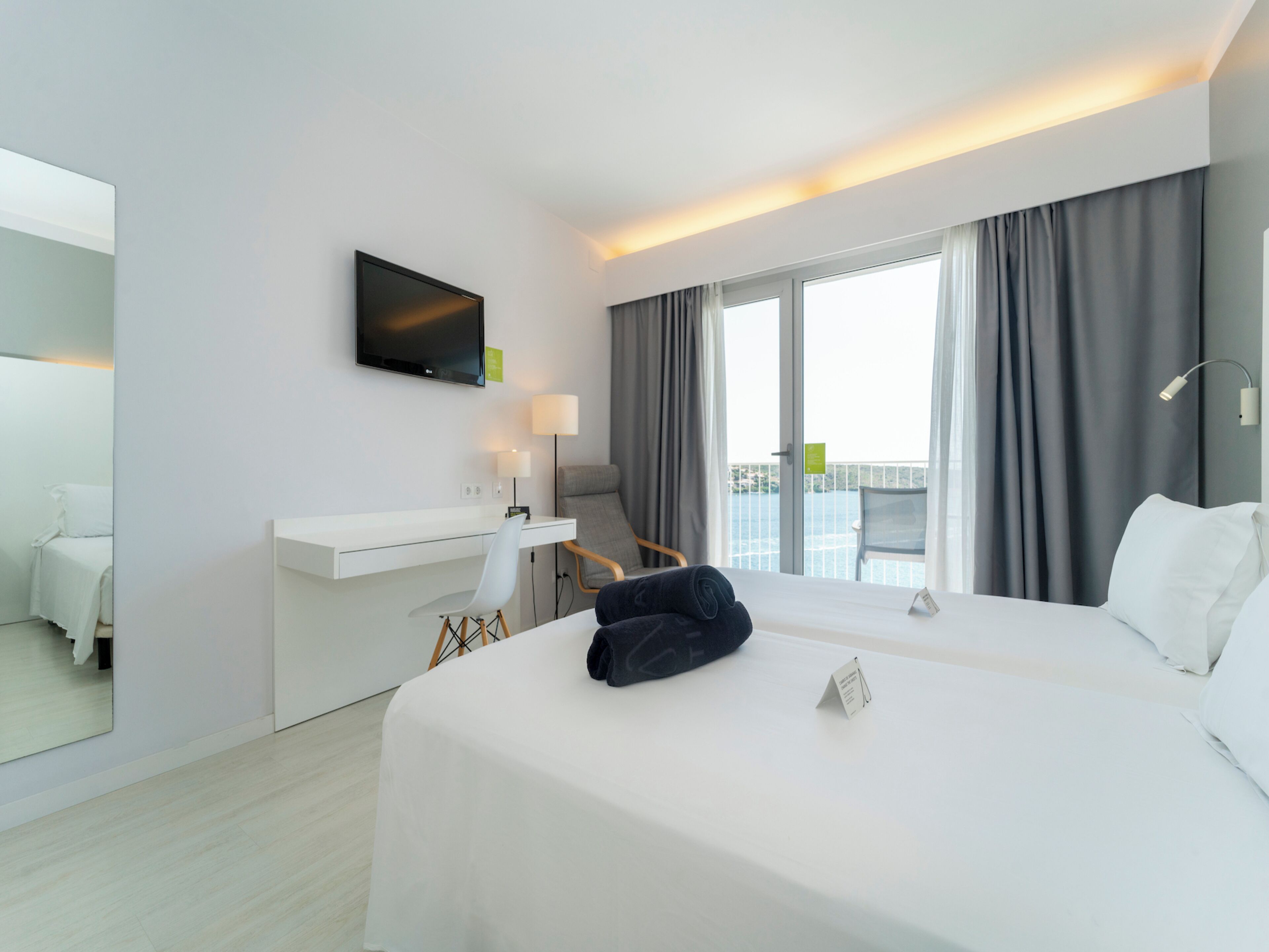 A modern hotel room with a large bed, a desk, a chair, a TV, and a balcony with curtains.