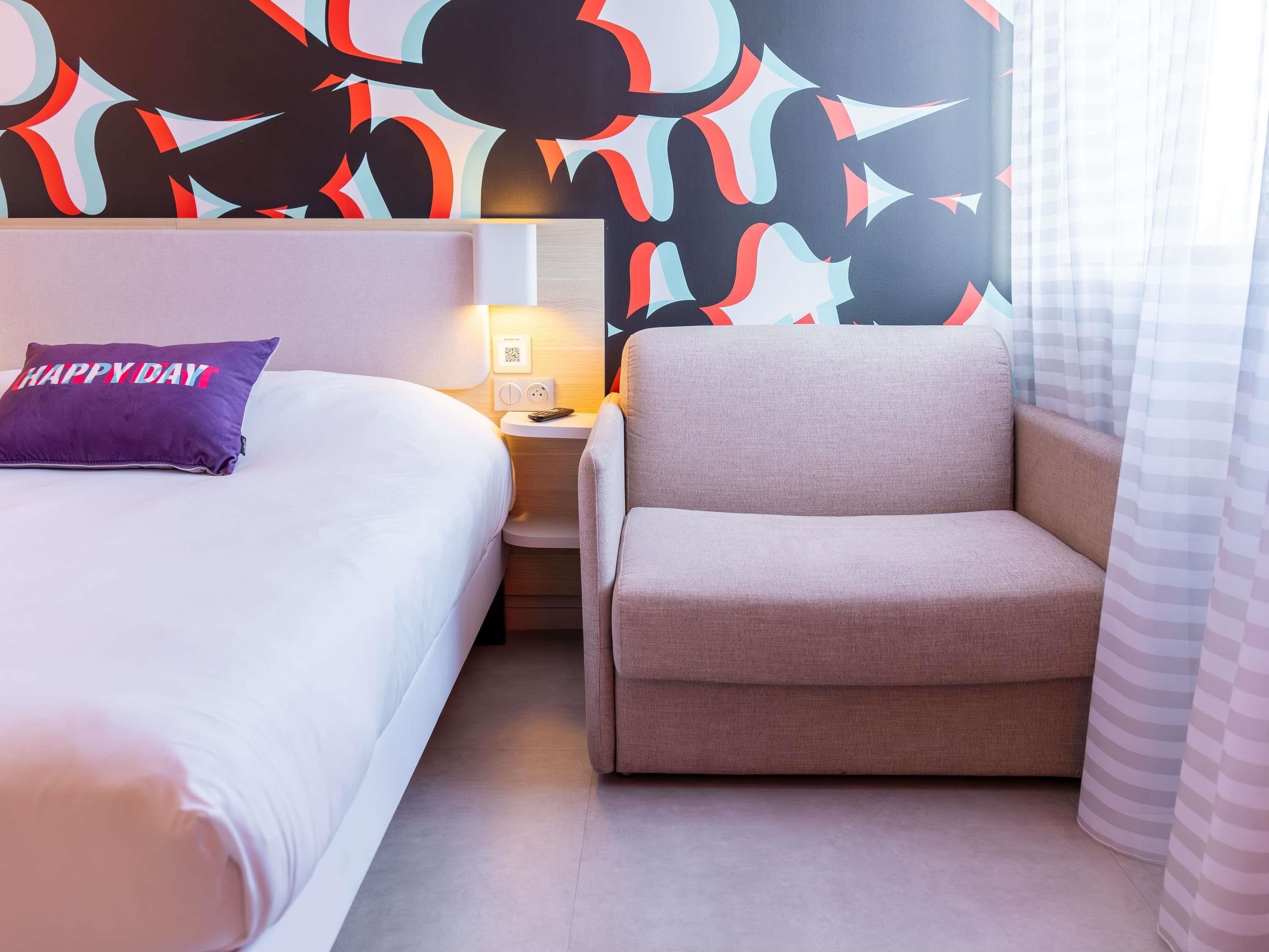 A hotel room with a bed, a chair, a nightstand, a lamp, and a wall with abstract art.