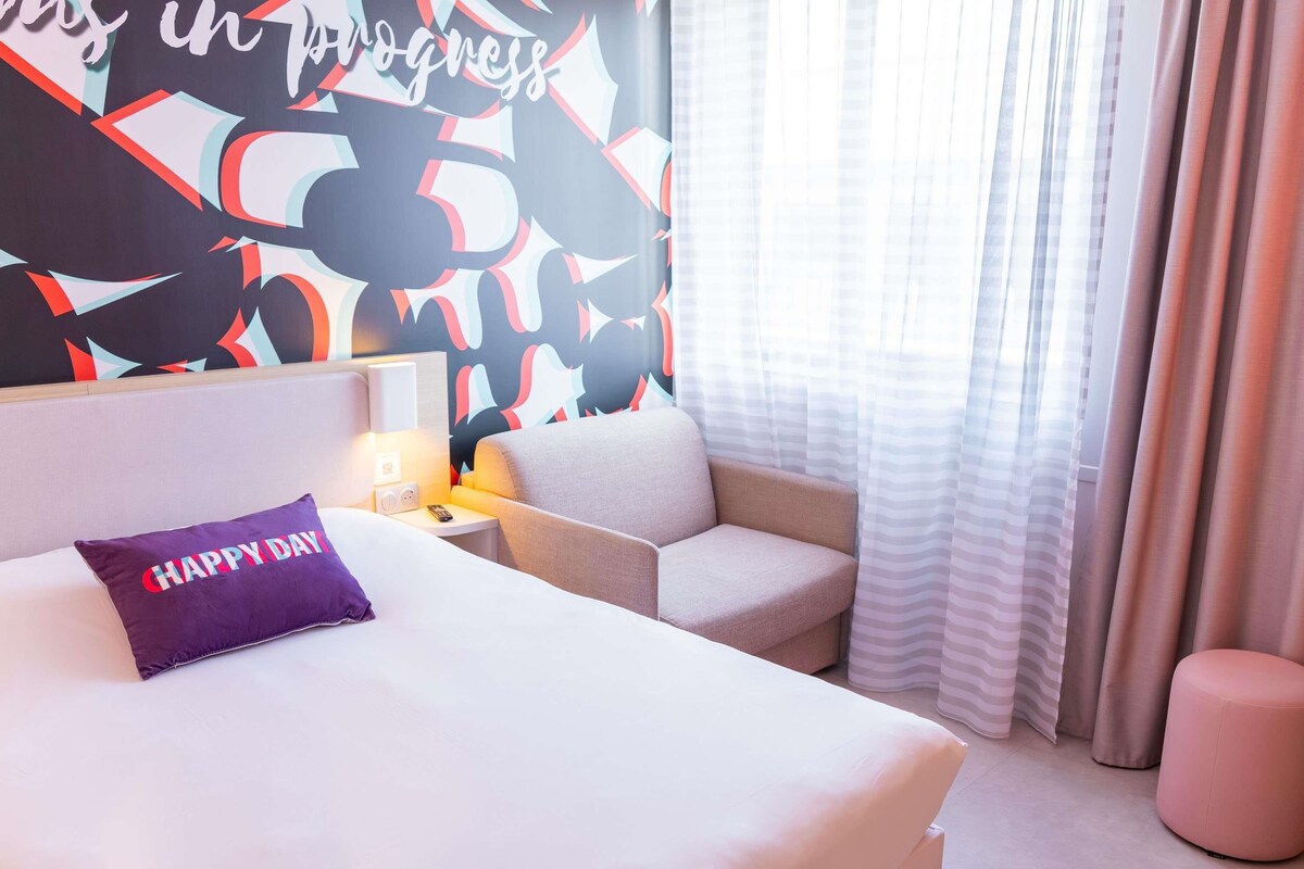 A hotel room with a bed, a chair, a nightstand, a lamp, and a wall with abstract patterns.