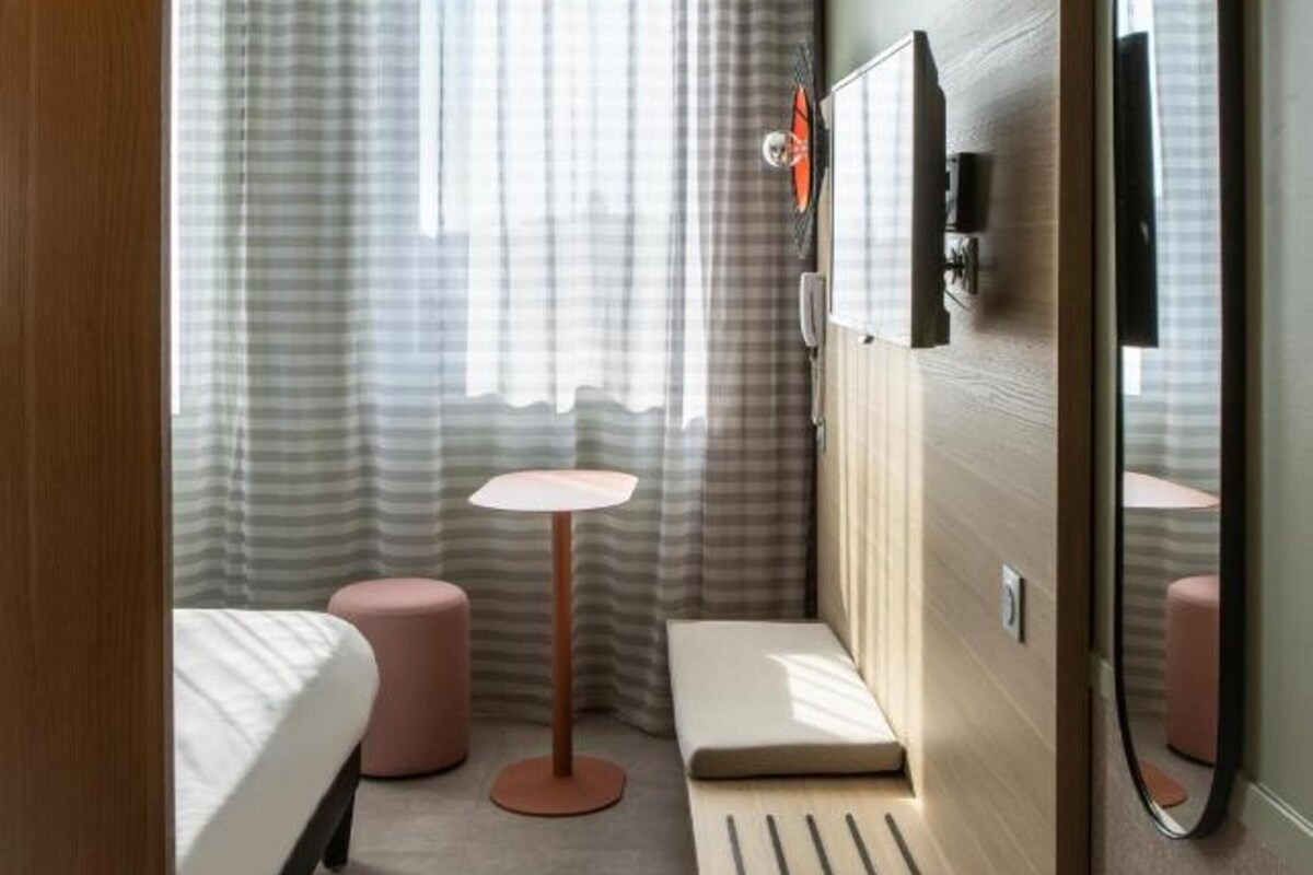 Ein kleines Hotelzimmer mit Bett, rundem Tisch, Bank, Spiegel und einem Fenster mit Vorhängen.