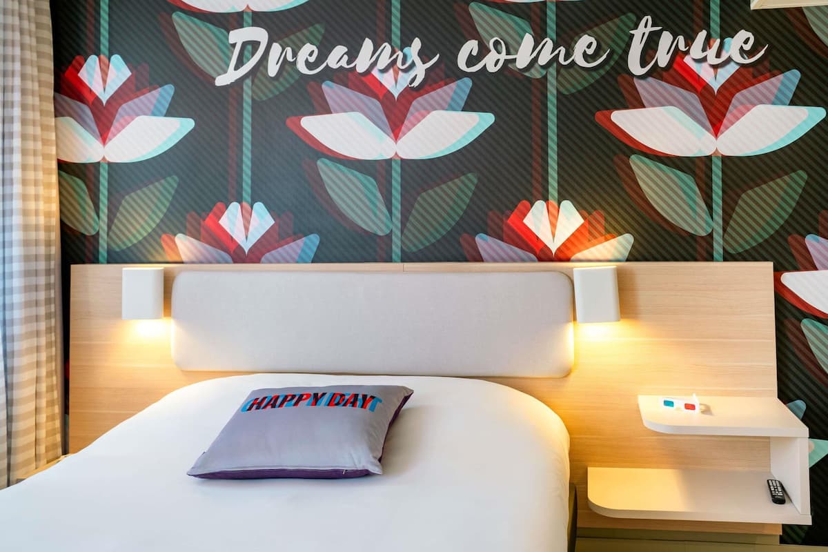 Habitación de hotel con una cama, un almohadón con el texto 'HAPPY DAY', una mesita de noche con un control remoto, y una pared con estampado floral y el texto 'Dreams come true'.