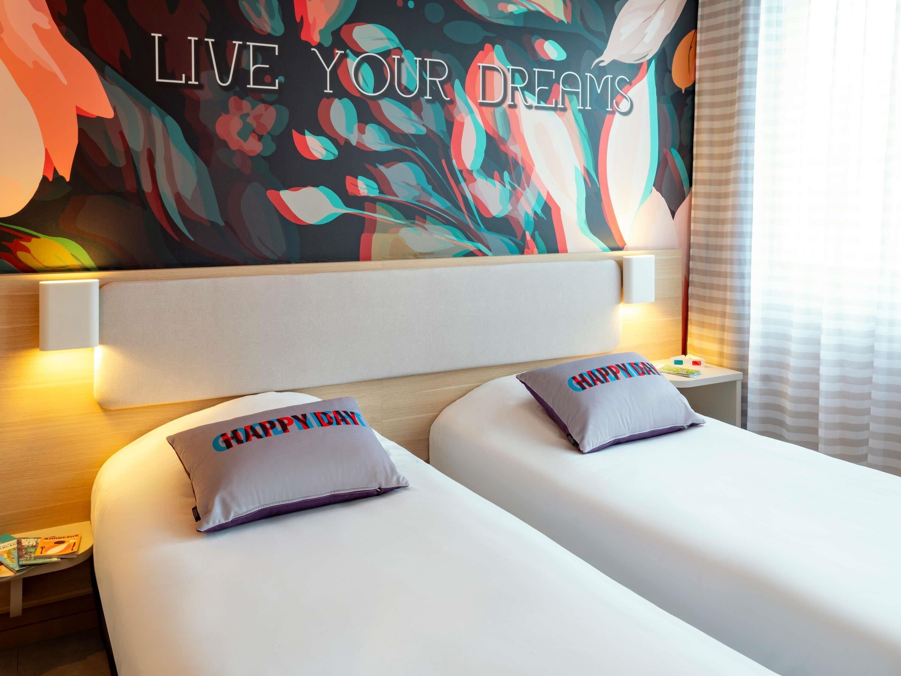 Una camera d'albergo con un letto, due cuscini, un comodino, una parete con una citazione motivazionale e una finestra con tende.