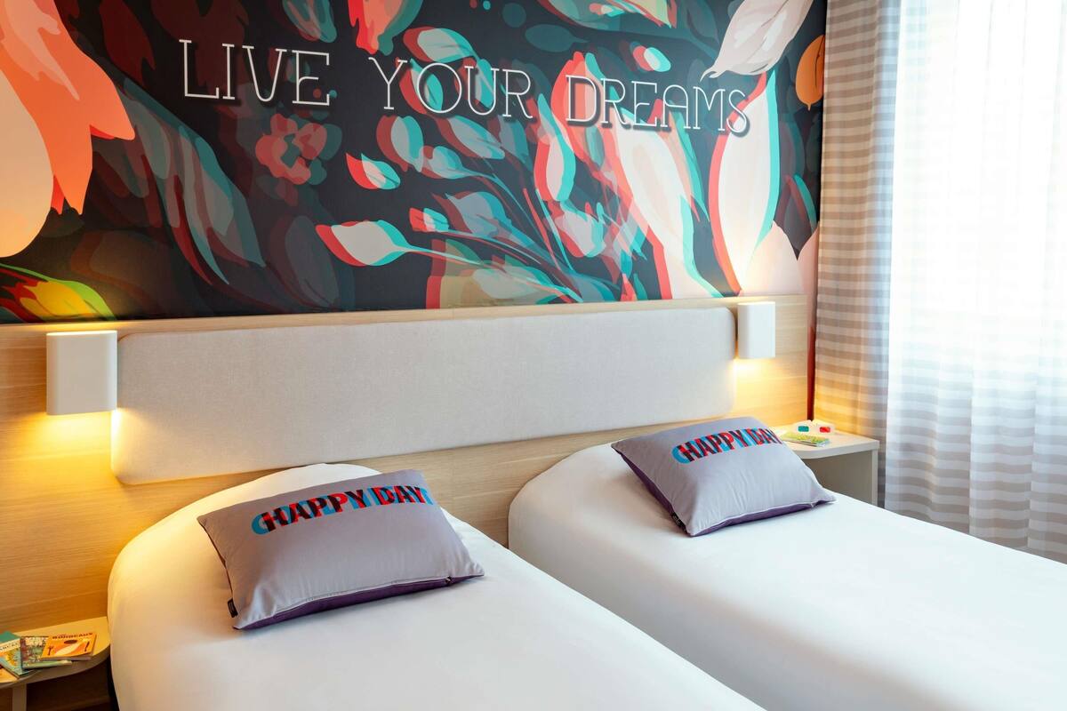 Habitación de hotel con cama, dos almohadas, mesita de noche, pared con una cita motivacional y ventana con cortinas.