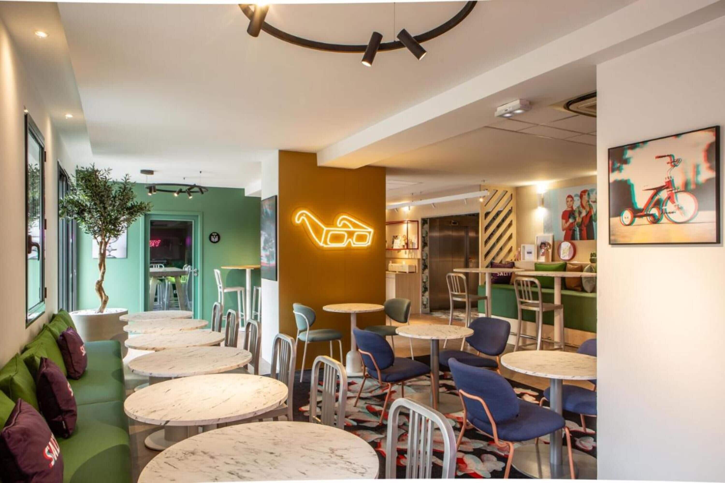 Un café moderno con mesas y sillas redondas, un letrero de neón y una pared verde.