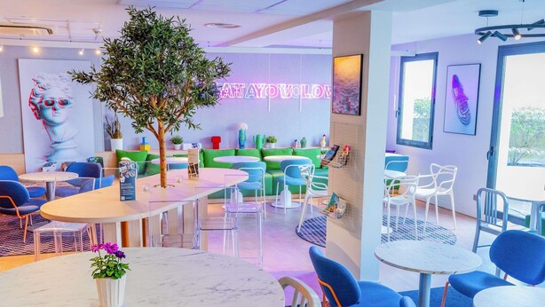 Een modern café met ronde tafels, blauwe stoelen en een neonbord.