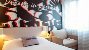 Een hotelkamer met een groot bed, een stoel, een nachtkastje en een muur met een kleurrijk abstract ontwerp.