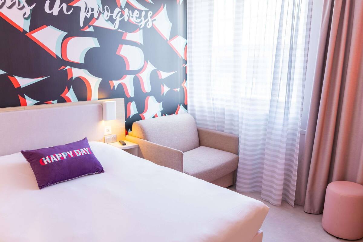 Habitación de hotel con cama, una silla, mesita de noche, lámpara y pared con diseños abstractos.