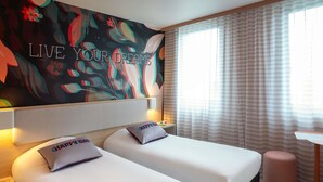 Habitación de hotel con dos camas, un cuadro grande con motivos florales en la pared, una ventana con cortinas y una mesita.