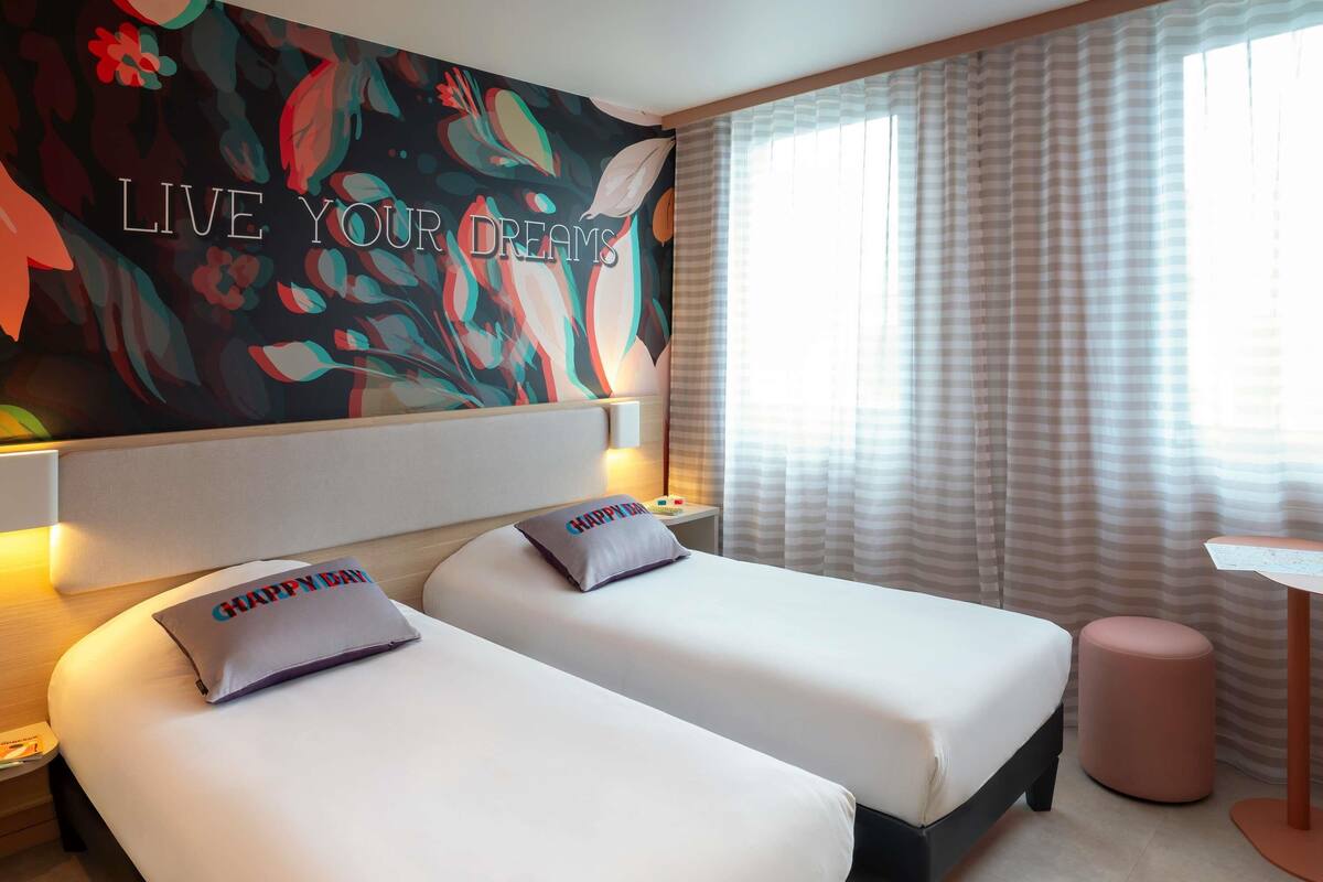 Habitación de hotel con dos camas, un cuadro grande con motivos florales en la pared, una ventana con cortinas y una mesita.