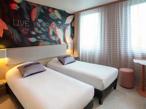 Habitación de hotel con dos camas, un cuadro grande con motivos florales en la pared, una ventana con cortinas y una mesita.