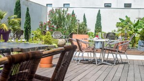 Un patio exterior con sillas y mesas de mimbre, rodeado de plantas en macetas y una pared blanca.