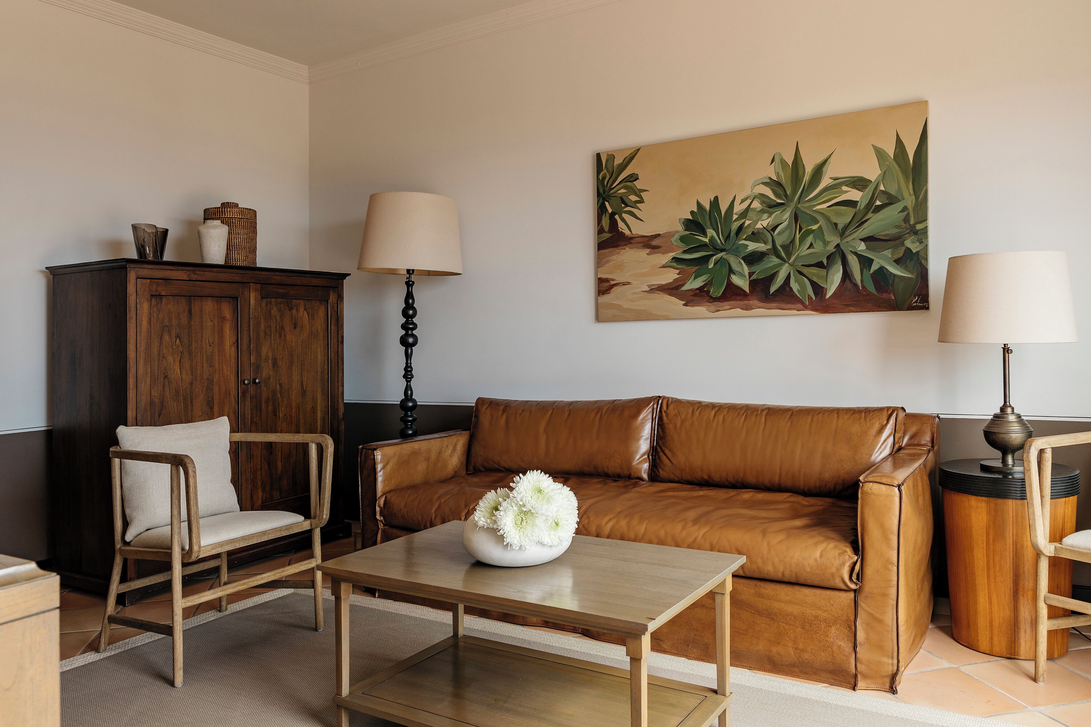A living room with a brown leather sofa, a wooden coffee table with a white vase, a dark wooden cabinet, and a painting of plants on the wall.