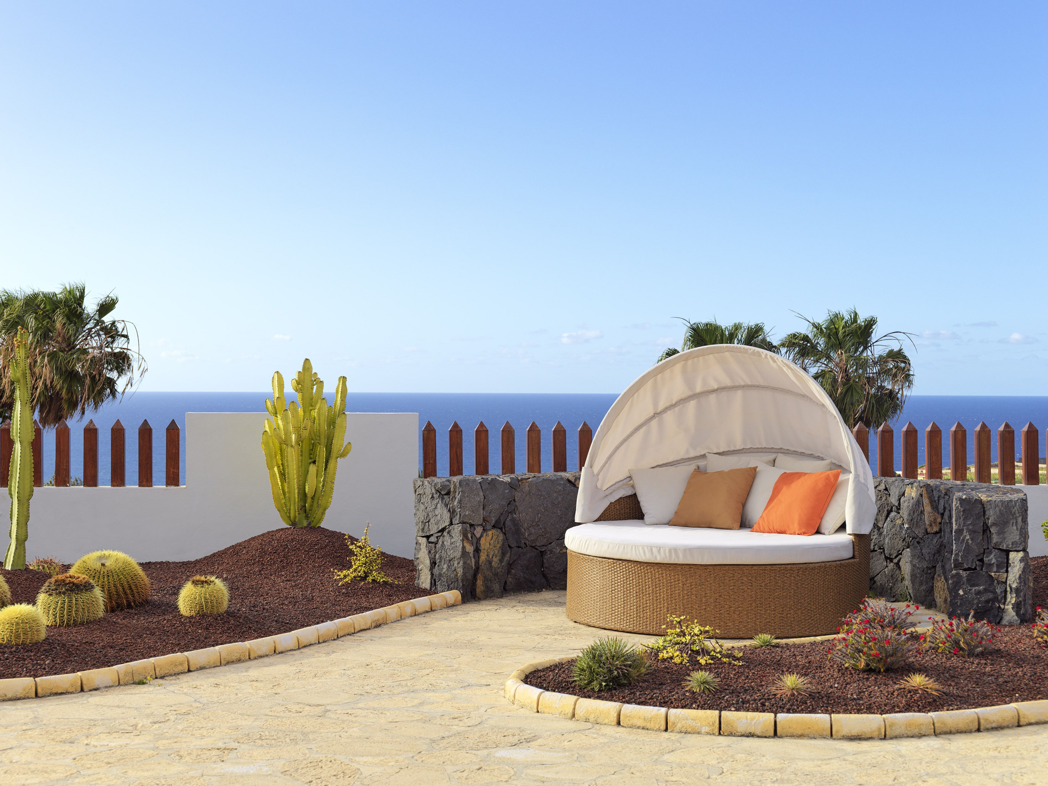 Een rond rieten bed met hemelbed, omringd door een tuin met cactussen en kleine planten, met uitzicht op de kust.