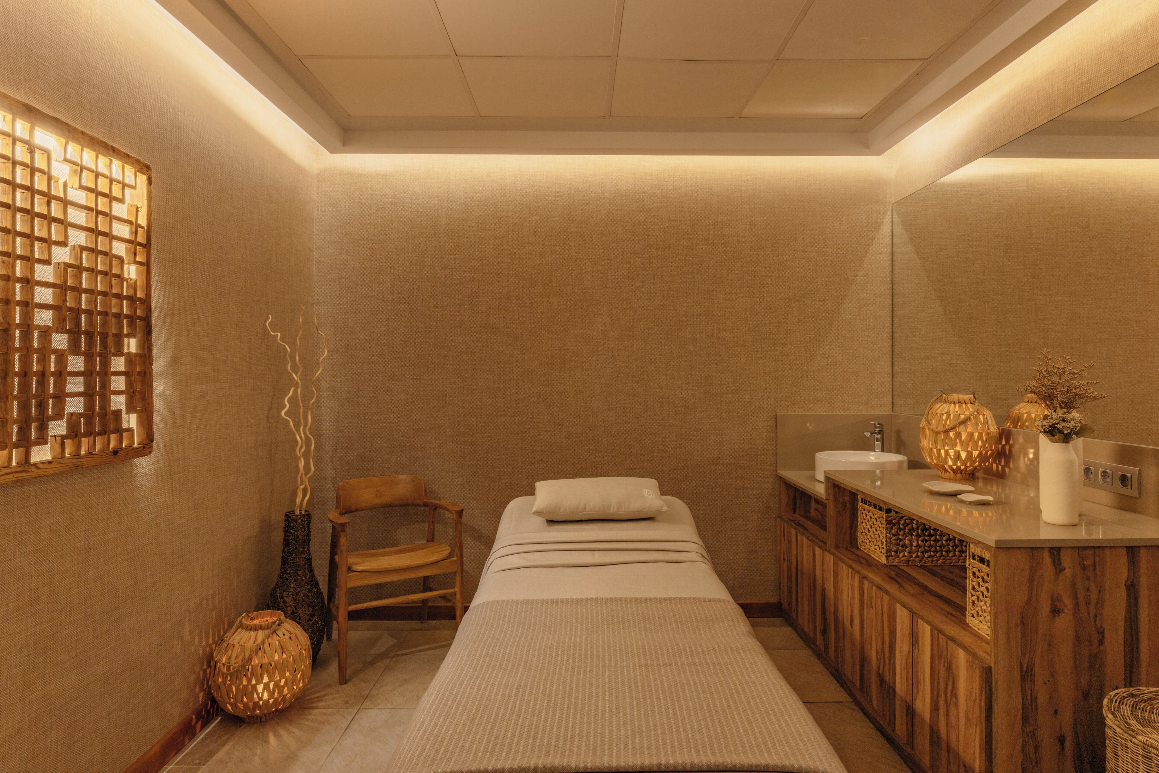 A room with a bed, a wooden chair, a small table, a mirror, and a sink.