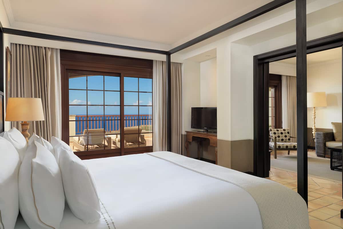 A hotel room with a large bed, a desk with a television, a balcony with a view of the sea, and a seating area with a chair and a table.