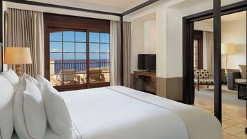 Habitación de hotel con una cama grande, un escritorio con televisor, un balcón con vistas al mar y una zona de estar con una silla y una mesa.