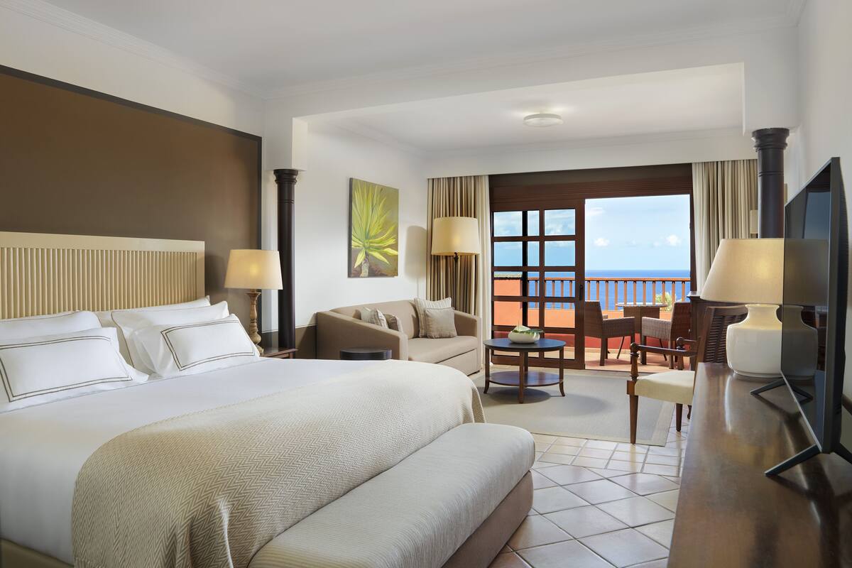 A hotel room with a large bed, a sofa, a dining table, and a balcony with a view of the ocean.