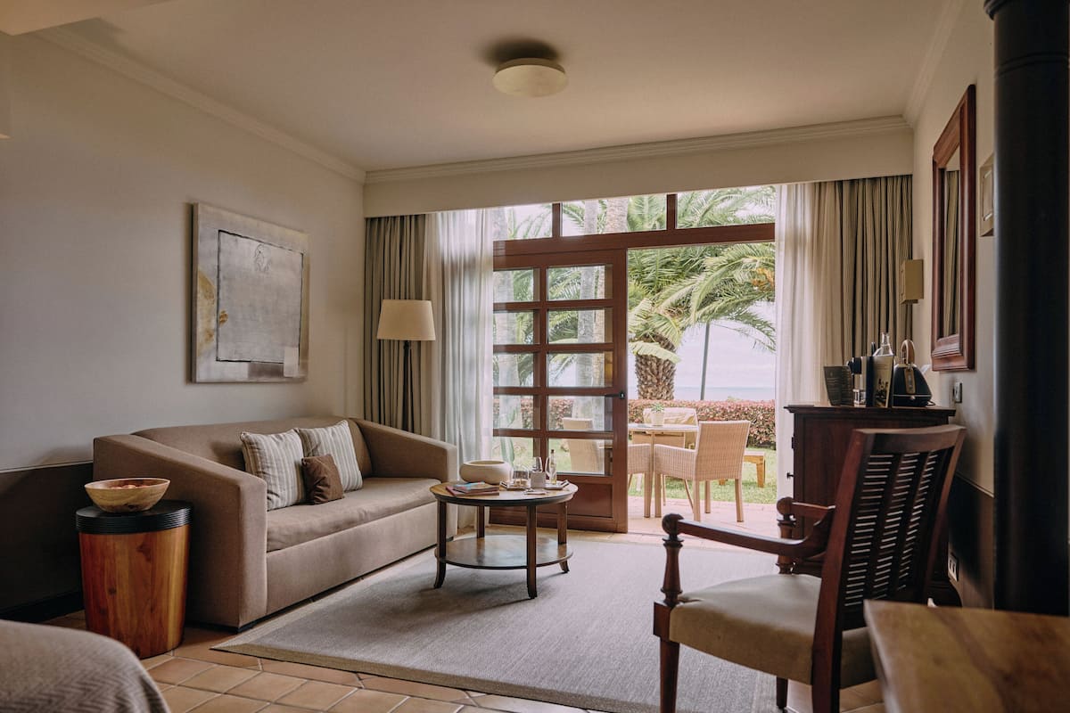 A living room with a sofa, a coffee table, and a chair. There is a window with curtains and a view of palm trees.