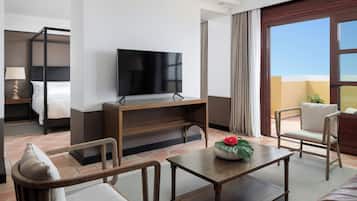 A hotel room with a bed, a TV, a coffee table, two chairs, a small table with a plant, and a view of the outdoors.