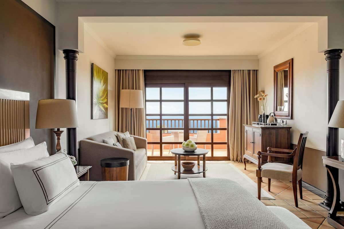 A hotel room with a large bed, a sofa, a small table with a vase of flowers, a desk with a lamp, and a view of the sea through the balcony doors.