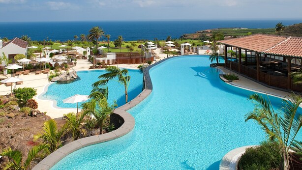 A resort pool with a curved design, surrounded by palm trees and lounge chairs, overlooking a coastal view. - Hotel Hacienda del Conde Meliá Collection Golf & Spa – Adults Only (Buenavista del Norte)