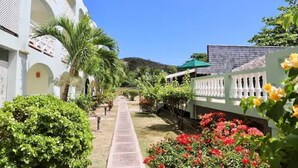 Un chemin menant à un bâtiment aux murs blancs et aux arcades, entouré de verdure et de plantes en fleurs.