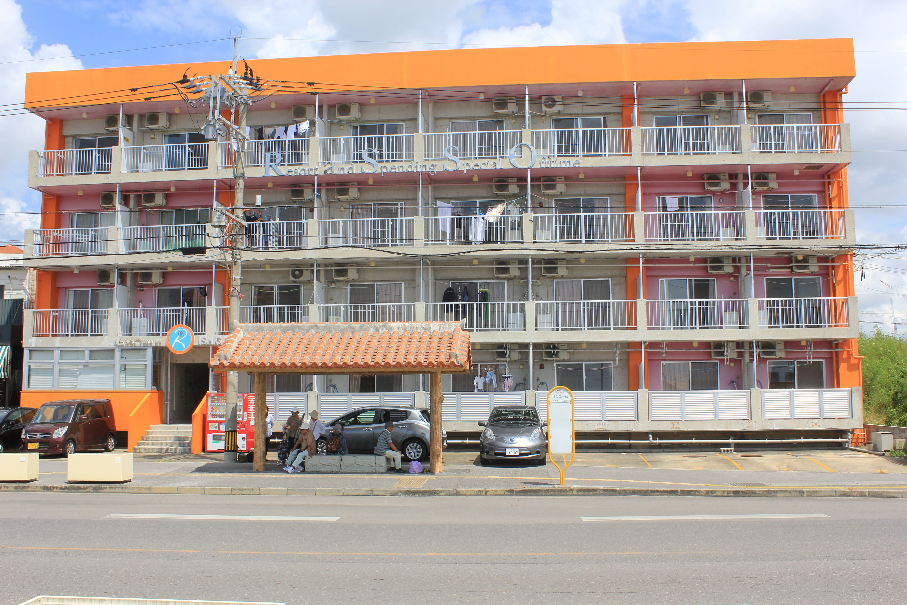 A multi-story building with balconies, a small covered area, and a parking lot.