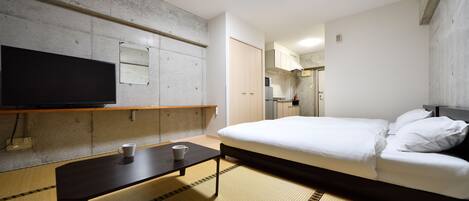 A hotel room with a bed, a flat-screen TV mounted on the wall, a small table with a cup, and a kitchenette area.