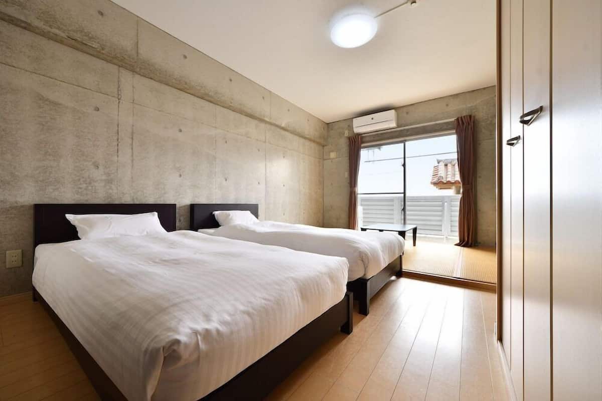 Une chambre avec deux lits, un mur en béton, une fenêtre avec des rideaux et un appareil de climatisation.