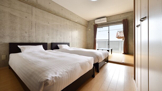 Un dormitorio con dos camas, una pared de concreto, piso de madera y una ventana con cortinas.