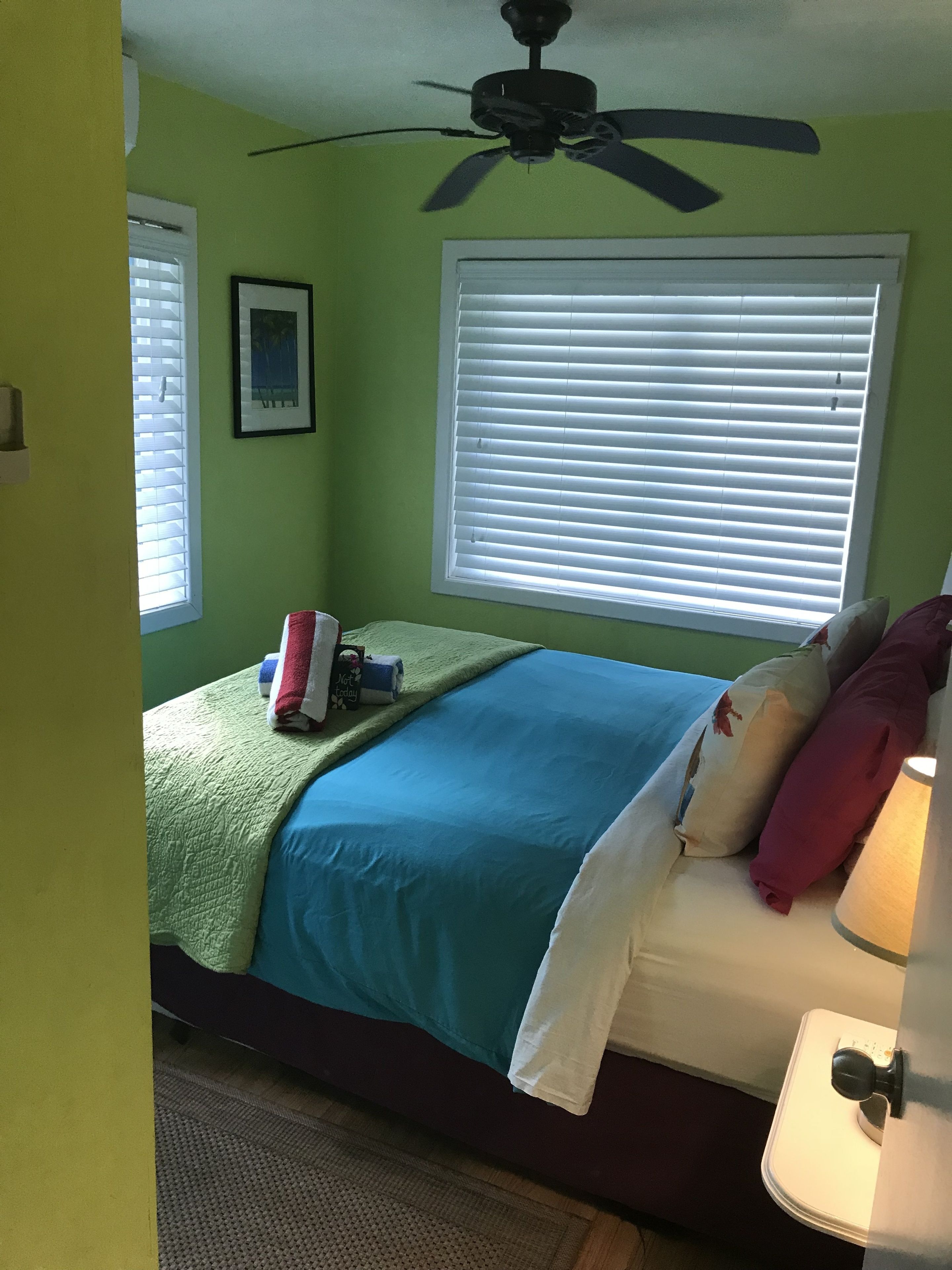 Een slaapkamer met een bed, een plafondventilator, een raam met zonwering en een bedlampje.