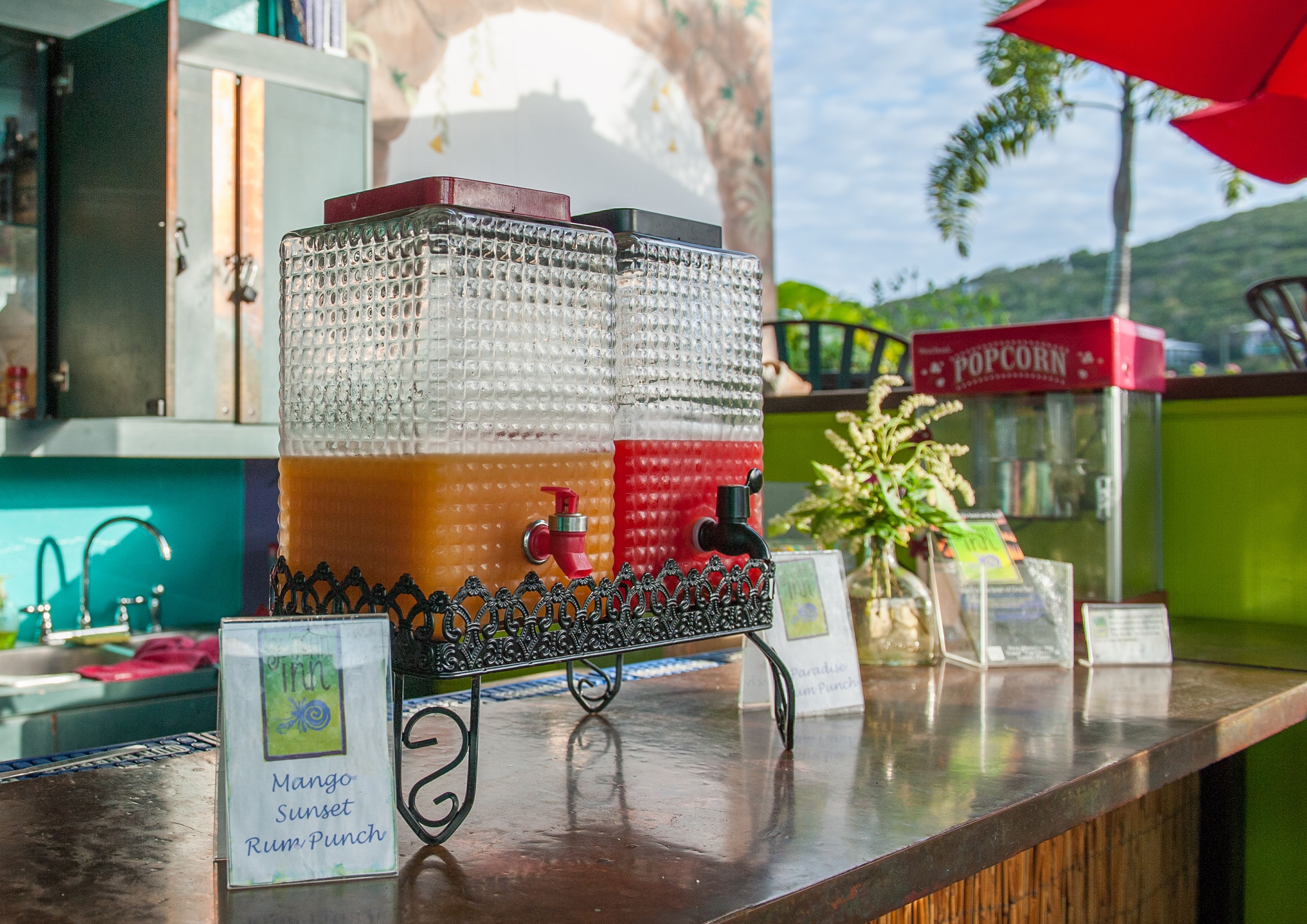 Una zona de bar con un gran recipiente de vidrio lleno de bebidas, una máquina para palomitas de maíz roja y un cartel anunciando el ponche con ron y mango, 'Mango Sunset'.